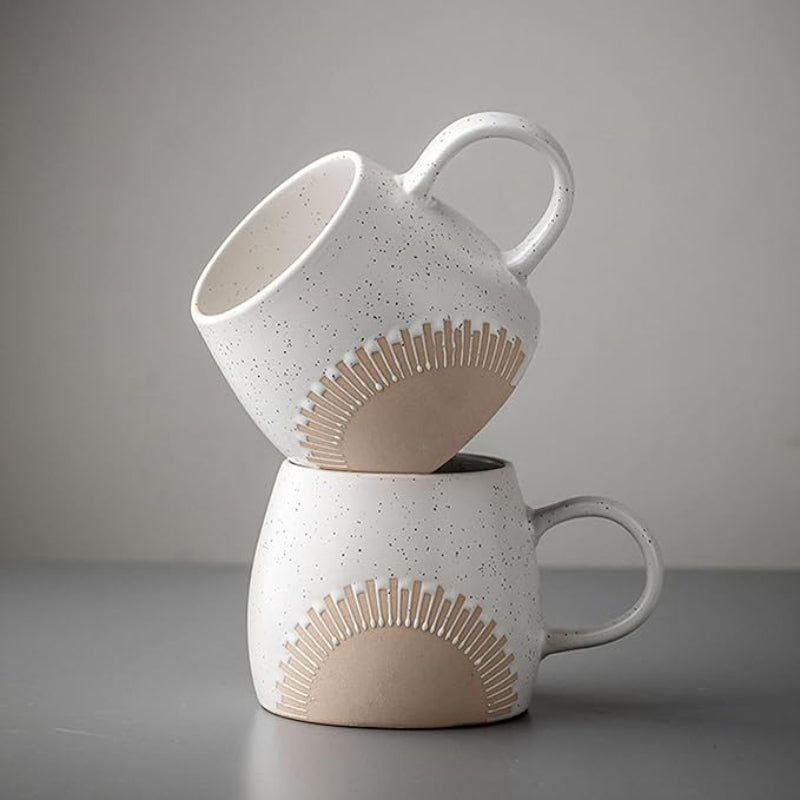 Two ceramic mugs with a speckled matte glaze and an embossed sunburst design are artistically stacked against a neutral gray background. The mugs have a curved handle and an unglazed base, showcasing a minimalist and modern aesthetic.