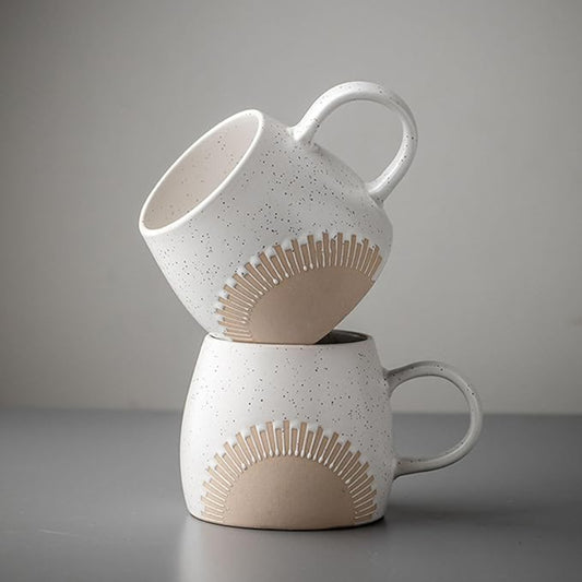 Two ceramic mugs with a speckled matte glaze and an embossed sunburst design are artistically stacked against a neutral gray background. The mugs have a curved handle and an unglazed base, showcasing a minimalist and modern aesthetic.