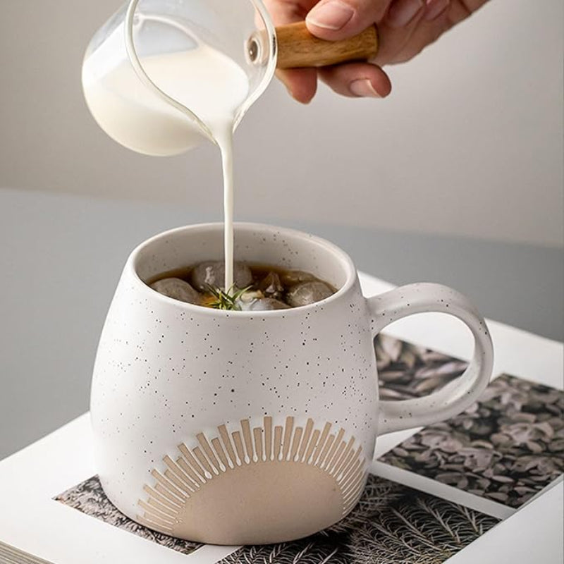 A ceramic mug with a speckled matte glaze and an embossed sunburst design. A hand is pouring milk from a glass pitcher into the mug, which contains iced coffee garnished with herbs. The mug has a curved handle and sits on an open magazine with nature-themed pages.