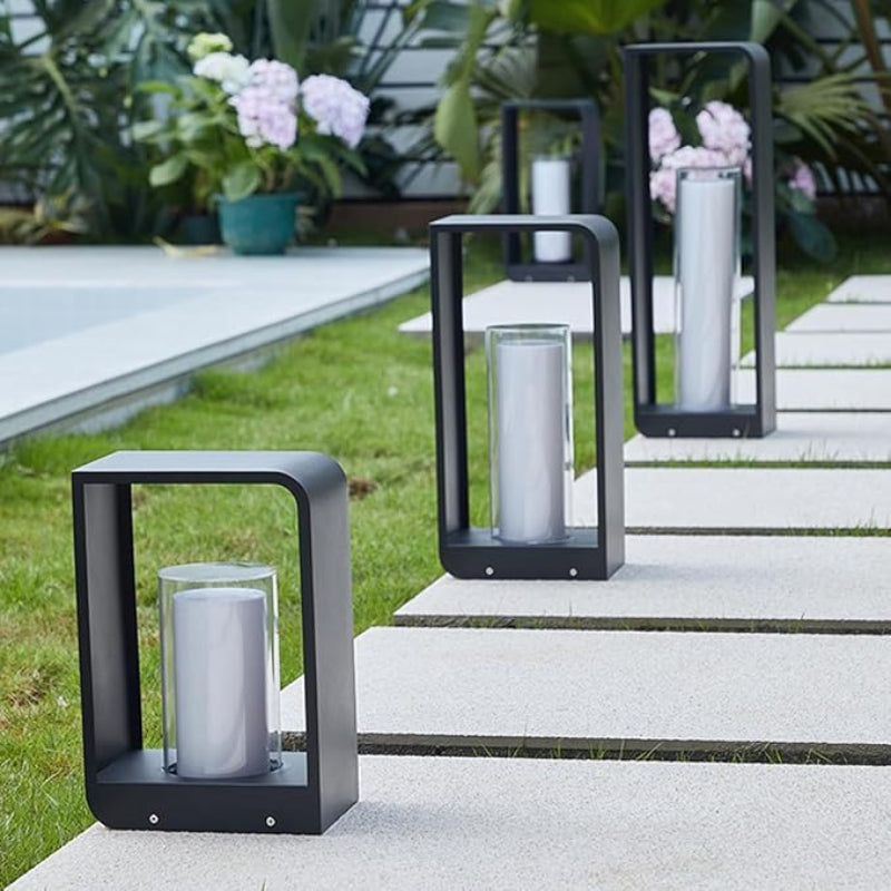 Three modern, dark rectangular outdoor pathway lanterns with frosted cylindrical diffusers, arranged along a stone paver path bordered by grass and flowering plants.