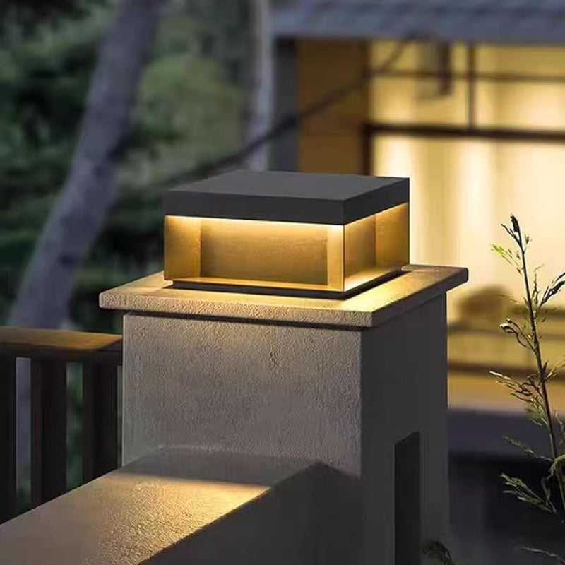 A modern, square outdoor light with a dark top and glowing warm-toned panels, mounted on a gray concrete pillar near a wooden railing at dusk.