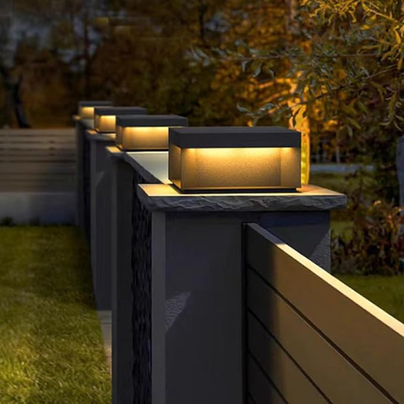 A series of modern, rectangular outdoor lights with dark tops and glowing warm-toned panels, mounted on top of dark gray fence posts along a walkway at dusk.