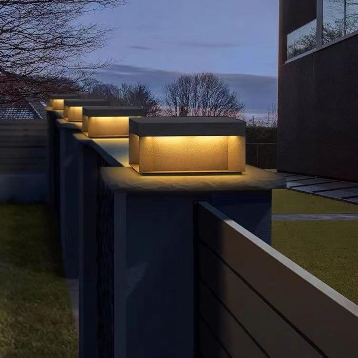 A row of modern, rectangular solar-powered lights emitting a warm glow, mounted on top of dark fence posts along a walkway at twilight.