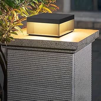 A modern, square outdoor light with a dark top and glowing warm-toned panels, mounted on a textured gray pillar near lush foliage at dusk.