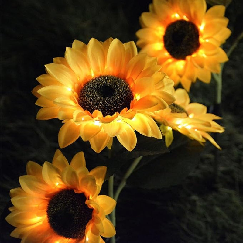Four illuminated artificial sunflowers with dark centers and warm yellow petals glow against a dark background at night. The petals are backlit, creating a bright outline around each flower.