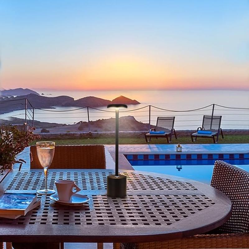 A cordless LED table lamp with a black cylindrical base, a slender stem, and a circular light panel emitting a warm glow, situated on a round outdoor patio table with a lattice design. The table also holds a wine glass, a white cup and saucer, and a book. In the background, a swimming pool with lounge chairs overlooks a scenic view of the ocean, islands, and a vibrant sunset sky.
