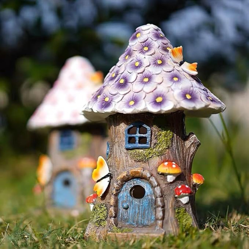 A small fairy house resembling a tree with a purple flower roof sits on white pebbles, featuring a blue door and window, and mushroom and butterfly decorations.