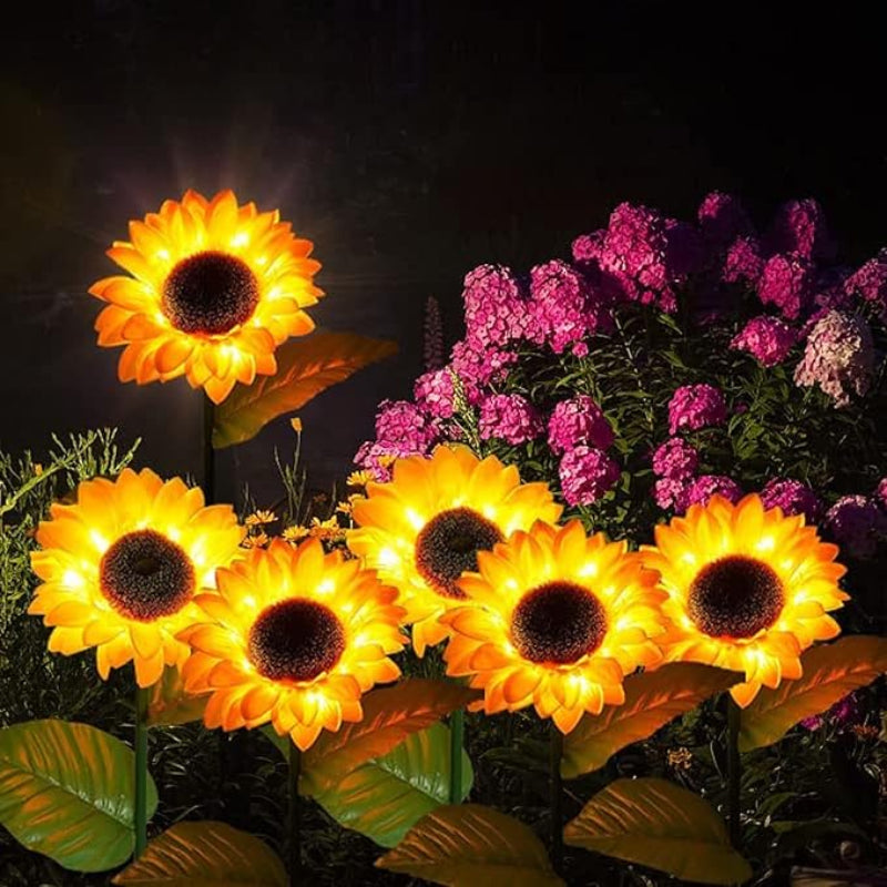 Several artificial sunflowers with dark centers and warm yellow glowing petals are arranged in a garden at night. The flowers are illuminated from within, creating a bright display against the dark background. Lush green leaves are visible at the base of the flowers, and a cluster of pink flowers is blurred in the upper right corner. The scene suggests a charming and illuminated garden decoration.