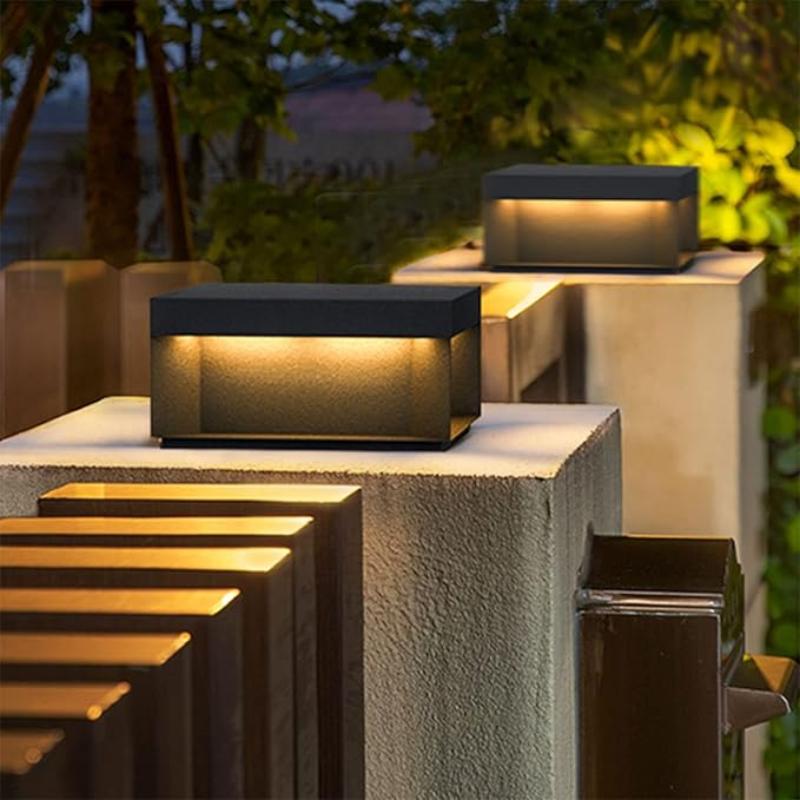 Two modern, rectangular outdoor lights with dark tops and glowing warm-toned panels, mounted on top of gray concrete pillars near wooden steps at dusk.