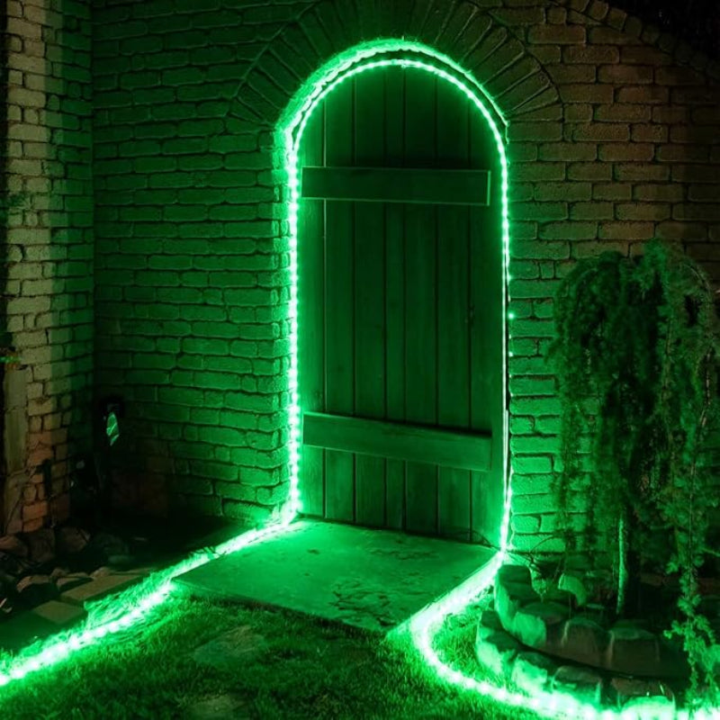 A vibrant green rope light outlines an arched doorway in a weathered brick wall at night. The green illumination also traces part of a stone pathway leading to the wooden door and the edge of a small, circular stone feature with a plant. The surrounding grass is also tinted green by the light.