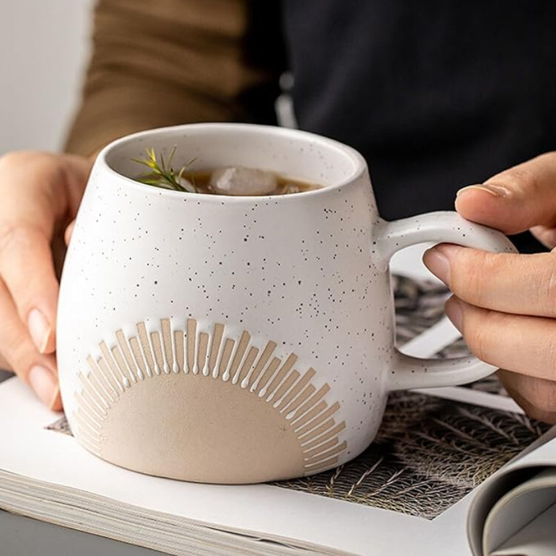 A person holding a speckled ceramic mug with an embossed sunburst design while resting it on an open book. The mug contains a beverage with ice cubes and a sprig of greenery, creating a cozy and refreshing scene.