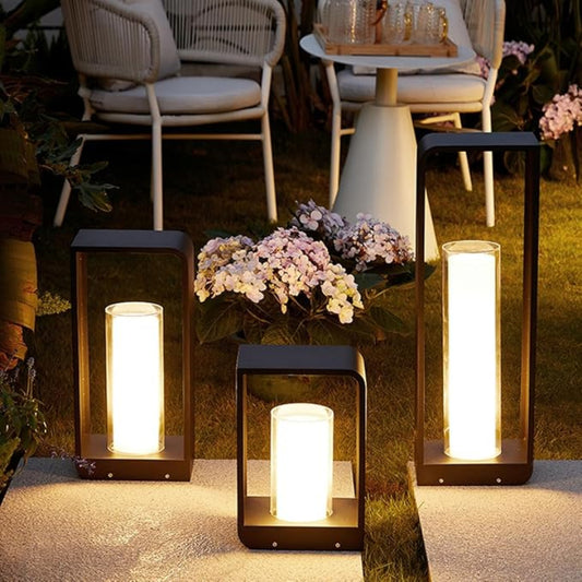 Three modern, dark-framed outdoor lanterns of varying heights with glowing cylindrical diffusers, illuminating a stone pathway and grassy area with outdoor seating and flowers at dusk.
