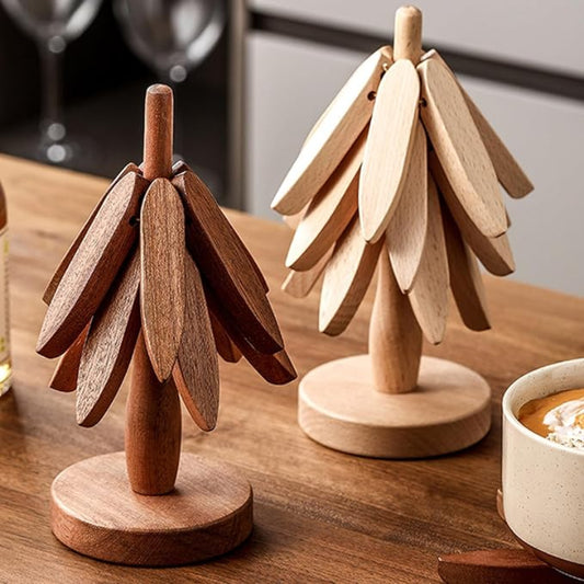 Two wooden trivets shaped like stylized trees, possibly "Tree Coasters," stand on a light wooden table. The one on the left is dark wood, and the one on the right is light wood. A mug is partially visible in the background.