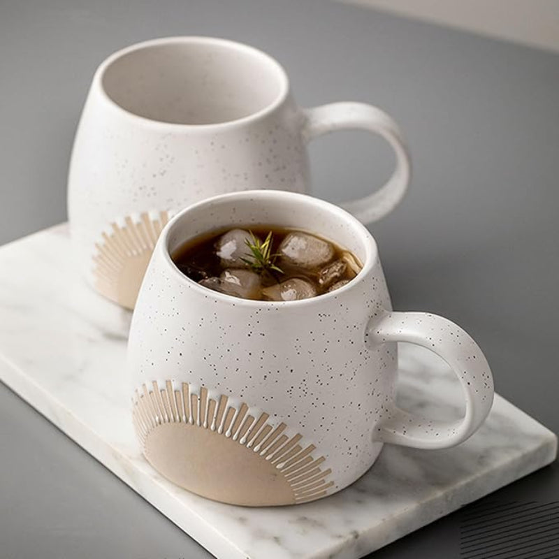 Two speckled ceramic mugs with an embossed sunburst design placed on a marble tray. One mug is filled with a cold beverage, ice cubes, and a sprig of greenery, creating a stylish and refreshing presentation.