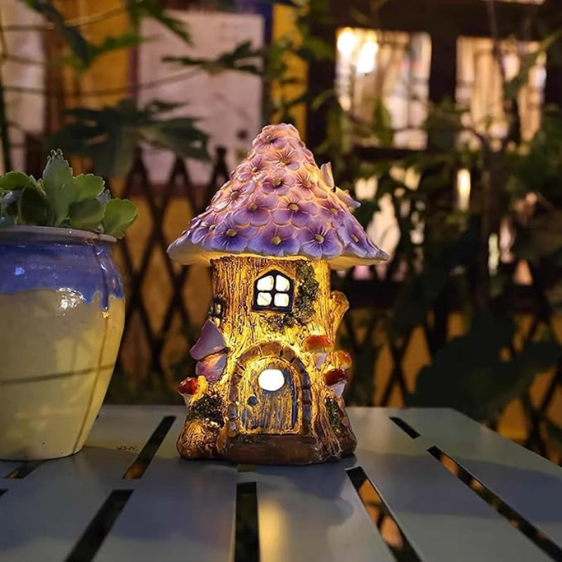 A small, illuminated fairy house with a purple flower roof sits on a slatted surface at night. It has a glowing arched doorway and window, and decorative mushroom and butterfly accents. A potted plant is to the left.