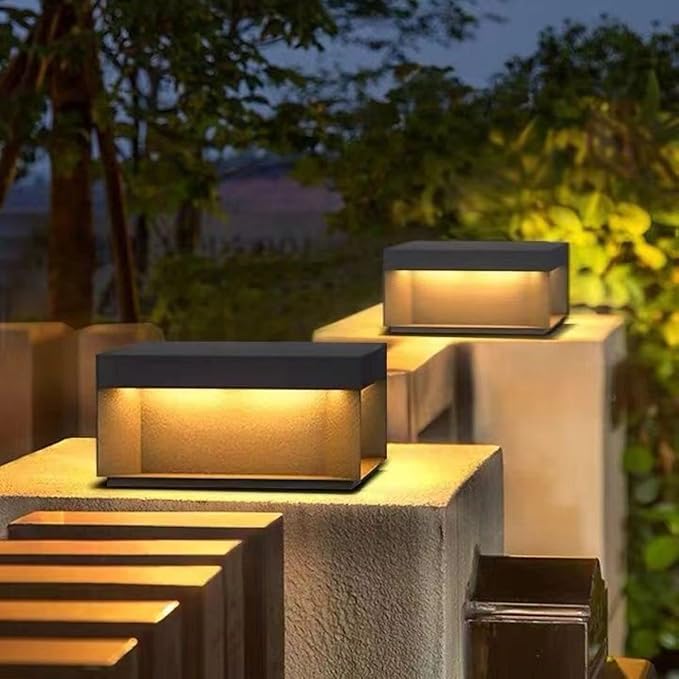 Two modern, rectangular outdoor lights with dark tops and glowing warm-toned panels, mounted on top of gray concrete pillars near wooden steps at dusk.