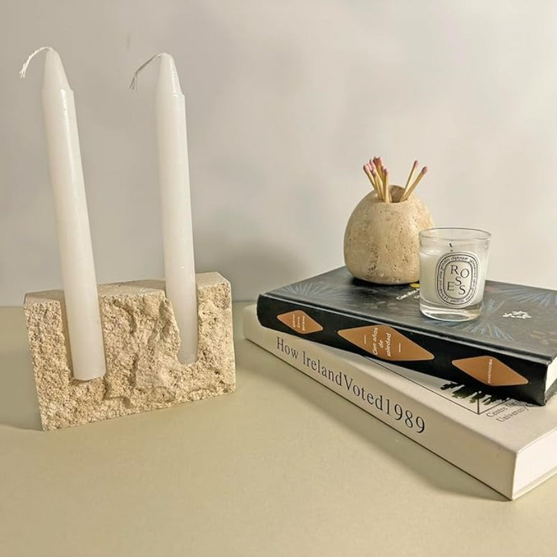 A textured, light-colored travertine stone block with two cylindrical openings holding two white taper candles, sitting on a light surface next to a stack of books topped with a small stone vase holding matches and a clear glass candle.