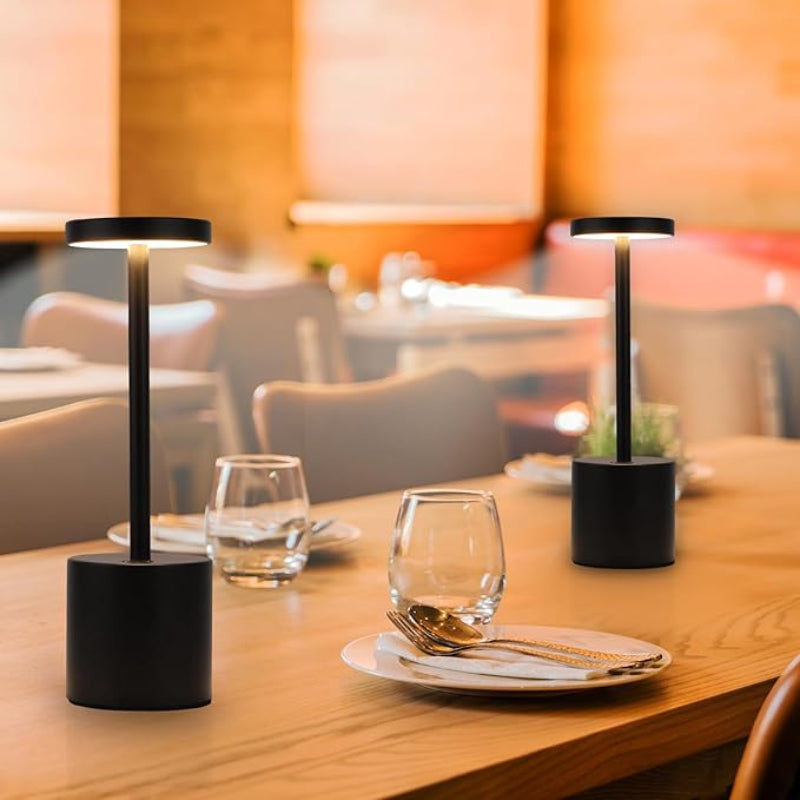 Two cordless LED table lamps with black cylindrical bases, slender stems, and circular light panels emitting a warm glow, are placed on a light wooden restaurant table. The table is also set with water glasses, plates, and gold-toned cutlery. The background shows a blurred restaurant setting with chairs and tables.