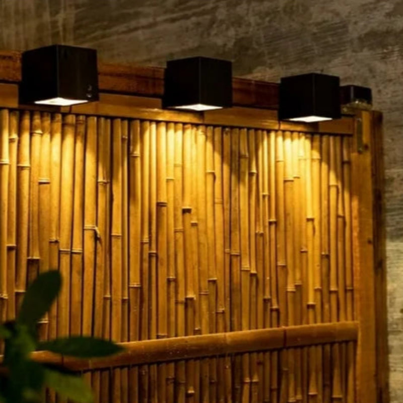 A row of modern black cube-shaped wall lights mounted on a wooden bamboo fence, casting warm downward illumination. The lights highlight the natural texture and vertical arrangement of the bamboo, creating a cozy and inviting ambiance. A portion of a green plant is visible in the foreground, adding a touch of nature to the setting.