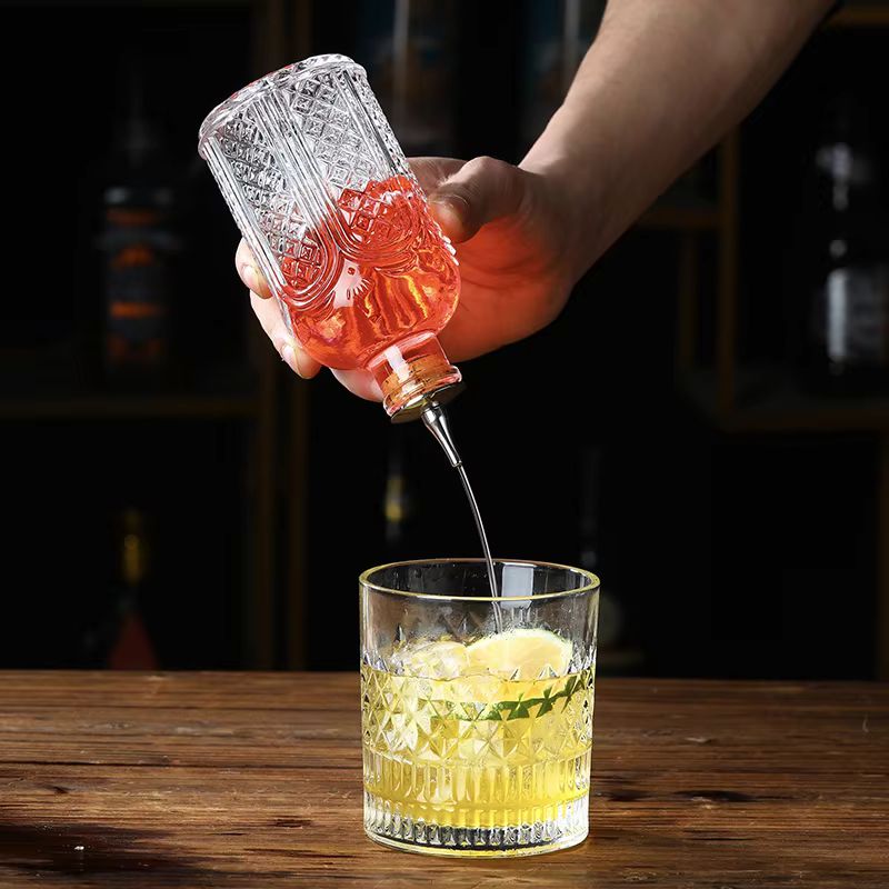 A hand pours red liquid from an intricately designed glass bitters bottle with a metal pour spout into a crystal-cut glass filled with ice and a yellow cocktail.