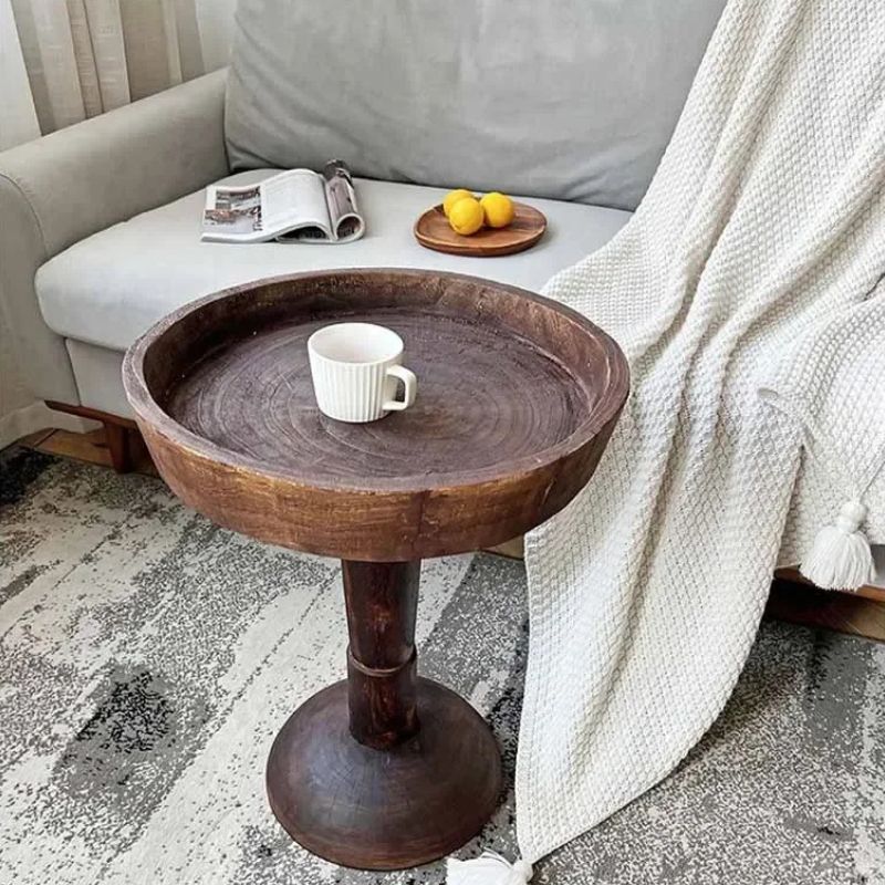 A rustic, dark wood, round side table (Momo Wabi-sabi Log Small Coffee Table) with a shallow tray top, holding a white cup, positioned next to a light-colored sofa with a magazine, lemons, and a white blanket.