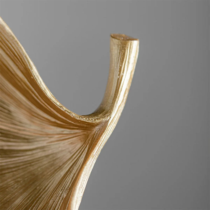 Close-up of a gold-colored decorative bowl featuring a curved, textured ginkgo leaf design against a neutral background.