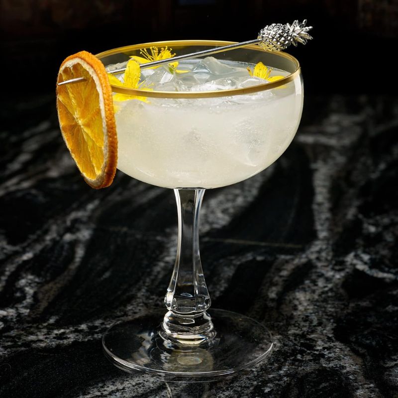 A cocktail in a gold-rimmed glass with a dried orange slice, yellow flowers, and a pineapple-shaped cocktail pick, on a dark marble surface.