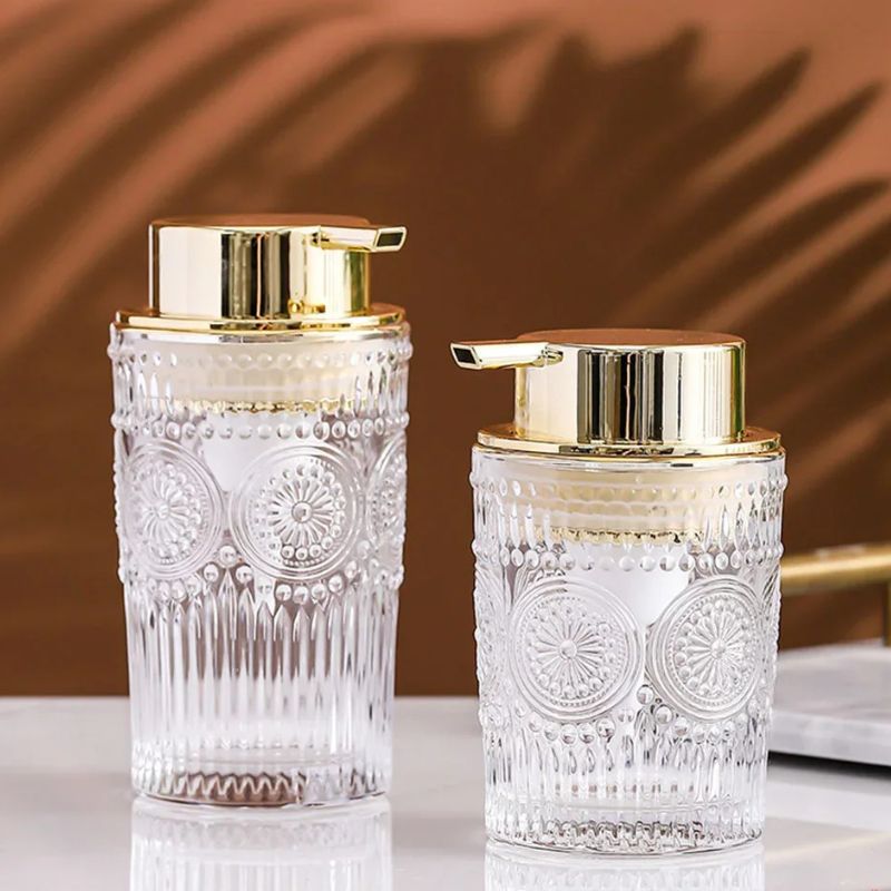 Two clear glass soap dispensers with ornate patterns and gold pumps, standing side-by-side on a light surface.