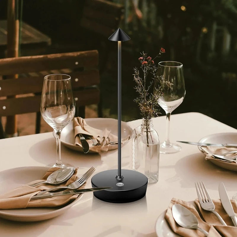 A cordless table lamp with a black, rounded base and a slim, vertical stem is shown on a table. The lamp has a pointed, conical shade. The table is set with plates, silverware, and wine glasses, suggesting a dining setting.