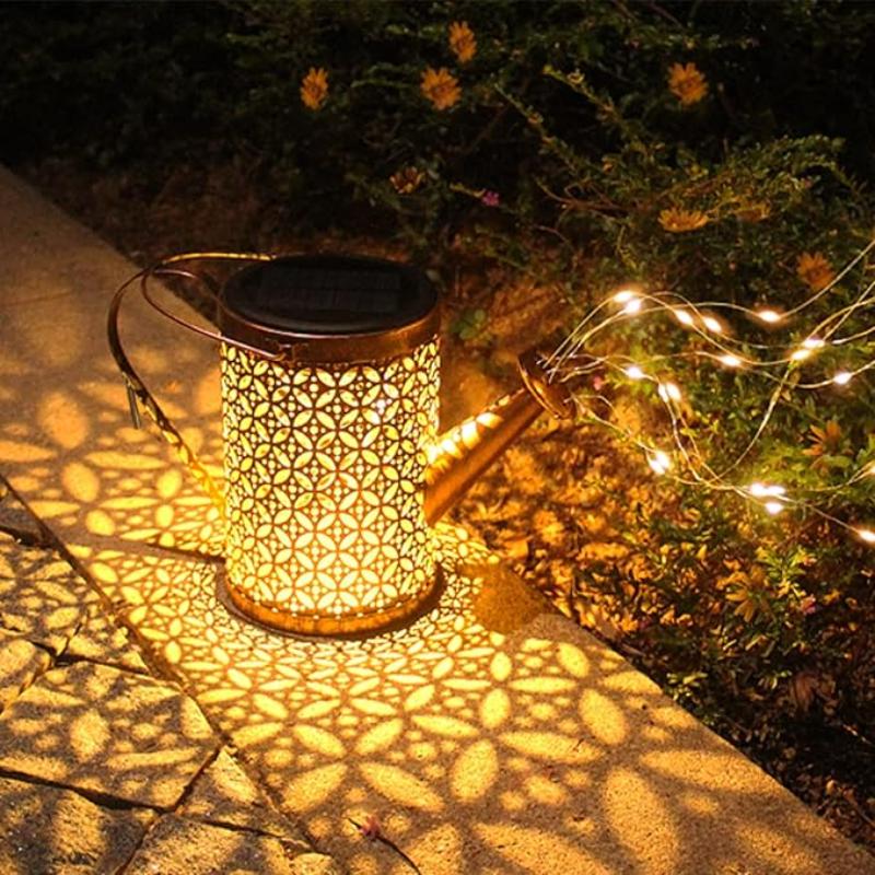 A decorative, bronze-toned watering can with intricate cutouts sits on a stone pathway at dusk or dawn. Solar-powered lights within illuminate the can and project floral patterns onto the ground. A stream of small, warm-toned LED lights extends from the spout, appearing to spill onto the adjacent greenery. The scene showcases an illuminated garden decoration.
