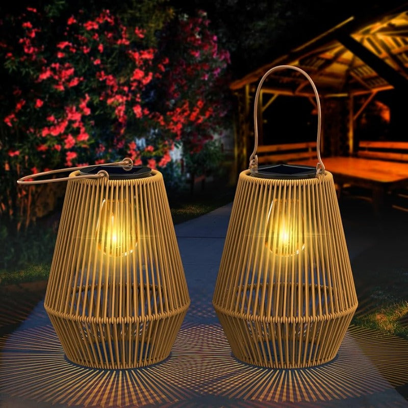 Two light brown, woven outdoor lanterns with vertical slats and dark solar panels on top emit a warm glow, casting radiating shadows on the ground at dusk or dawn. Each lantern has a metal handle for hanging. Blurred foliage and a wooden structure are visible in the background.