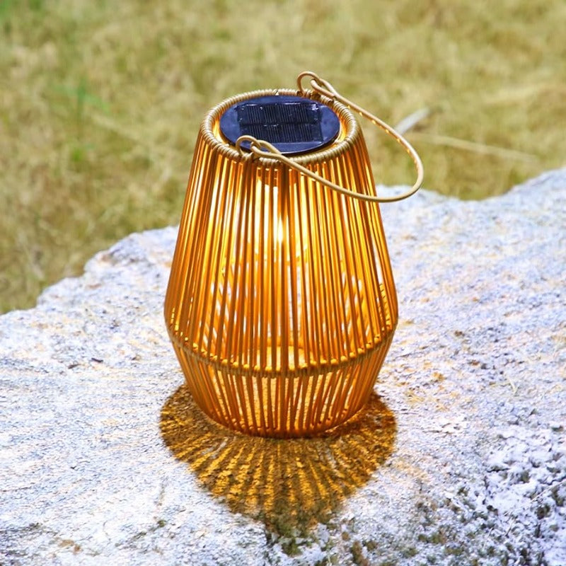 A warm-toned, woven outdoor lantern with vertical slats and a dark solar panel on top sits on a textured light gray stone surface. The lantern emits a soft glow and has a wire handle. Blurred green foliage is visible in the background.