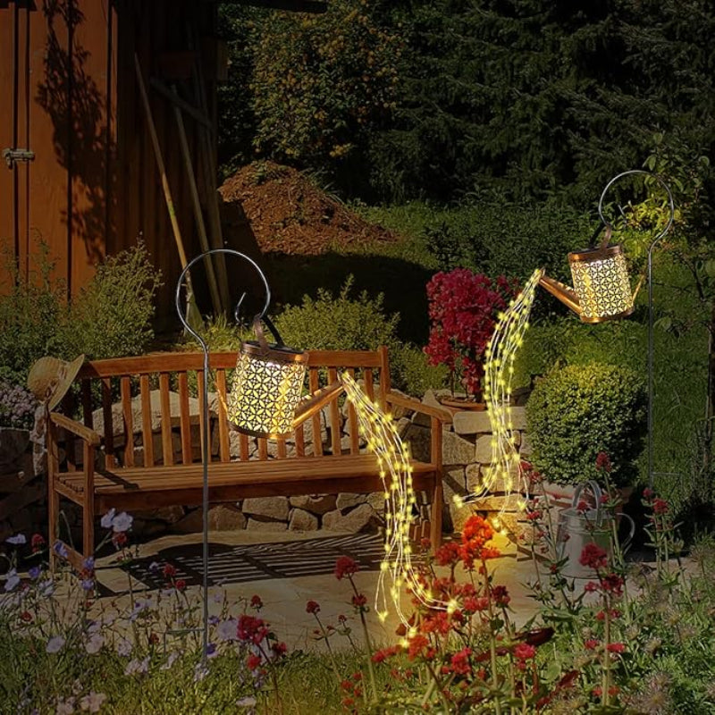 Two decorative, golden watering cans with intricate cutouts hang from black shepherd's hooks in a garden at dusk. Solar-powered lights within each watering can illuminate, casting patterns of light and creating a stream of small, warm-toned lights that appear to pour onto the surrounding flowers and greenery. A wooden bench sits in the background, and the overall scene evokes a magical, illuminated garden display.