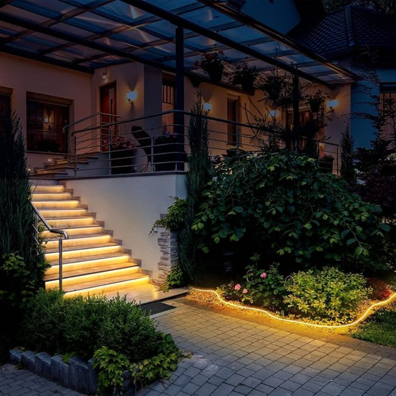 Warm white rope lights illuminate the edges of outdoor steps with metal railings leading up to a house at dusk. The rope lights also outline a paved walkway through a garden with lush greenery. The house has a glass-covered porch with hanging plants and soft lighting.