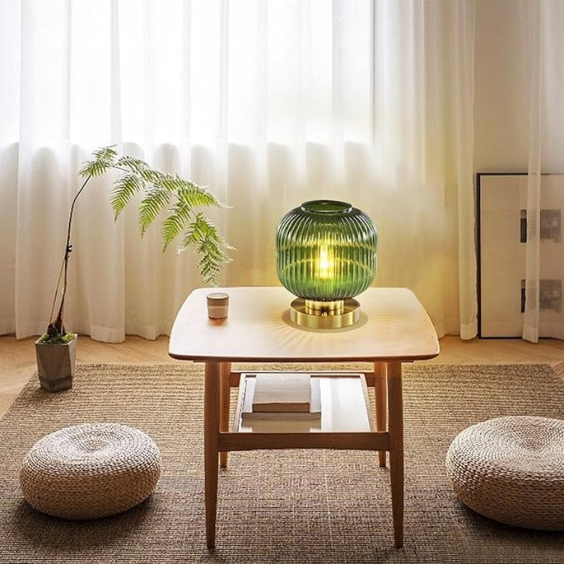 Lit green glass lamp on a small table in a bright room with cushions and a plant.