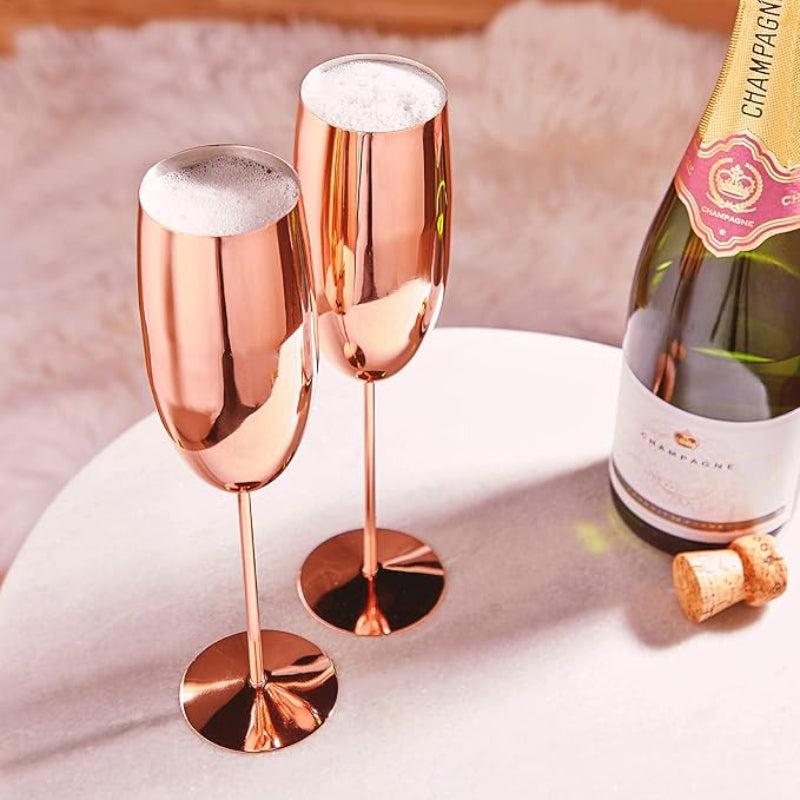 Two elegant, copper-toned stemmed glasses are filled with sparkling wine, topped with a white foam. They stand on a round white table next to a partially visible bottle of champagne and a champagne cork. The background is a soft, light-colored rug.