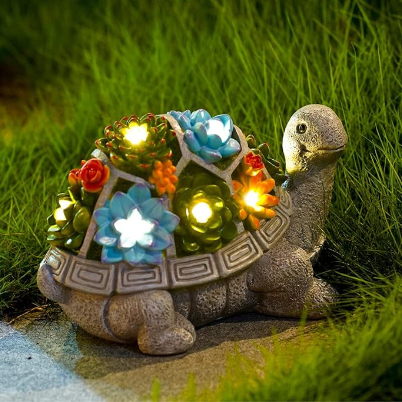 A gray decorative turtle statue with a segmented shell holding illuminated colorful artificial succulents is placed outdoors on a stone surface surrounded by green grass at dusk or night. The lights within the succulents emit a soft, warm glow.