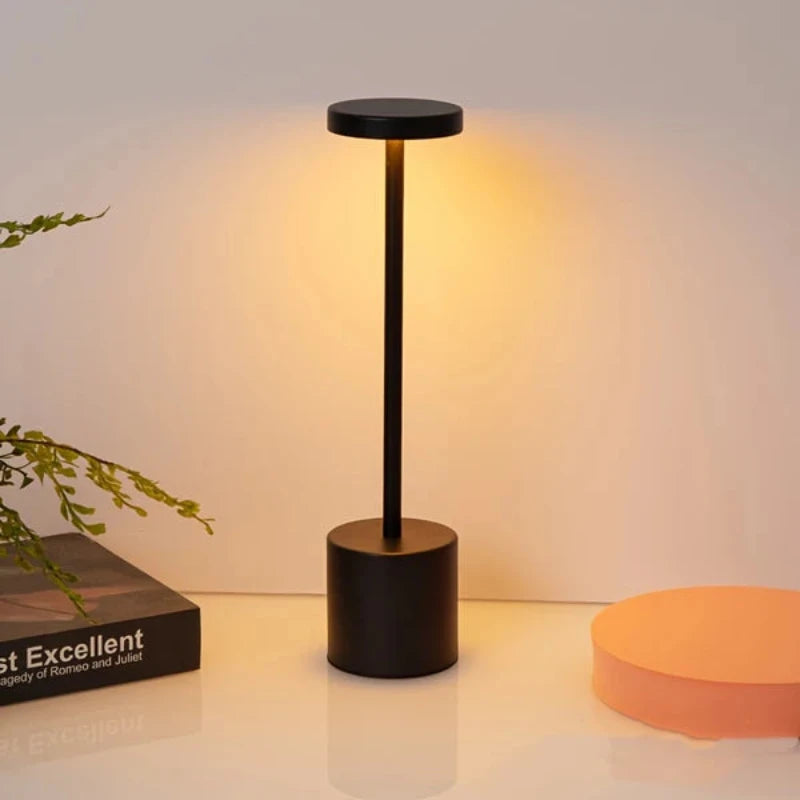 A cordless, rechargeable LED table lamp with a brushed gold finish, featuring a cylindrical base, a slender stem, and a circular light panel emitting a warm glow, positioned on a light-colored surface next to a book and a round orange object.