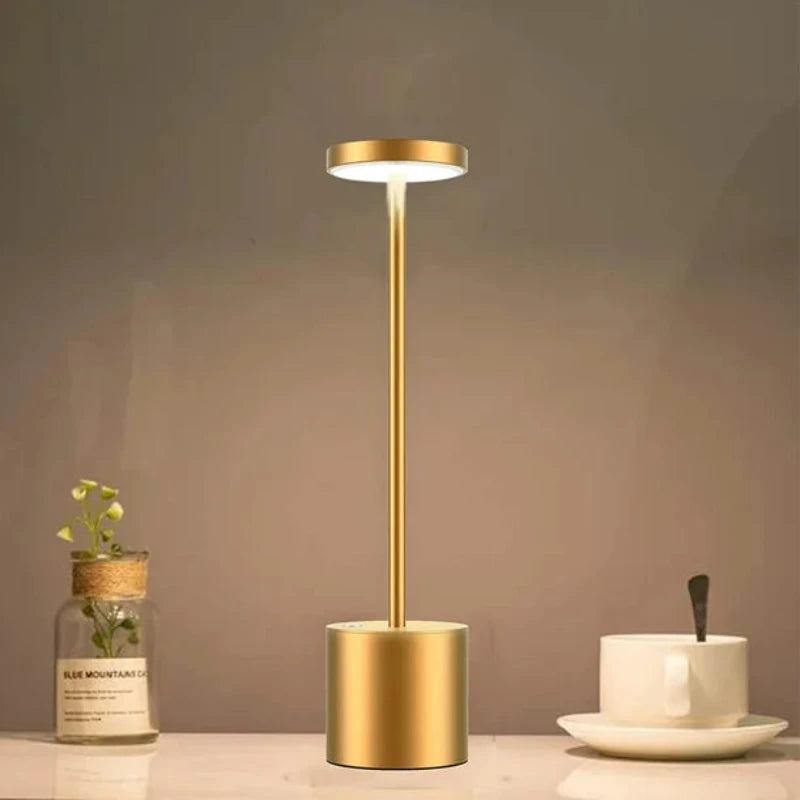A cordless LED table lamp with a brushed gold finish, featuring a cylindrical base, a slender stem, and a circular light panel emitting a bright white light, positioned on a light-colored surface next to a small glass jar with artificial greenery and a white cup and saucer with a spoon. The background is a warm beige.