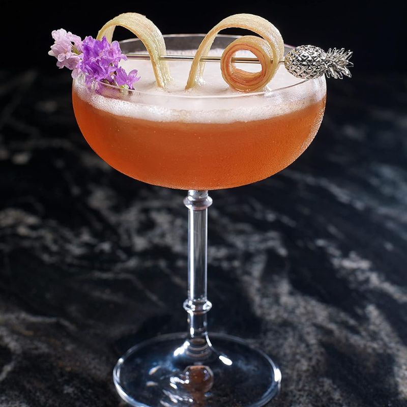 A pink cocktail in a stemmed glass, garnished with flowers, citrus peel, and a pineapple-shaped cocktail pick, on a dark marble surface.