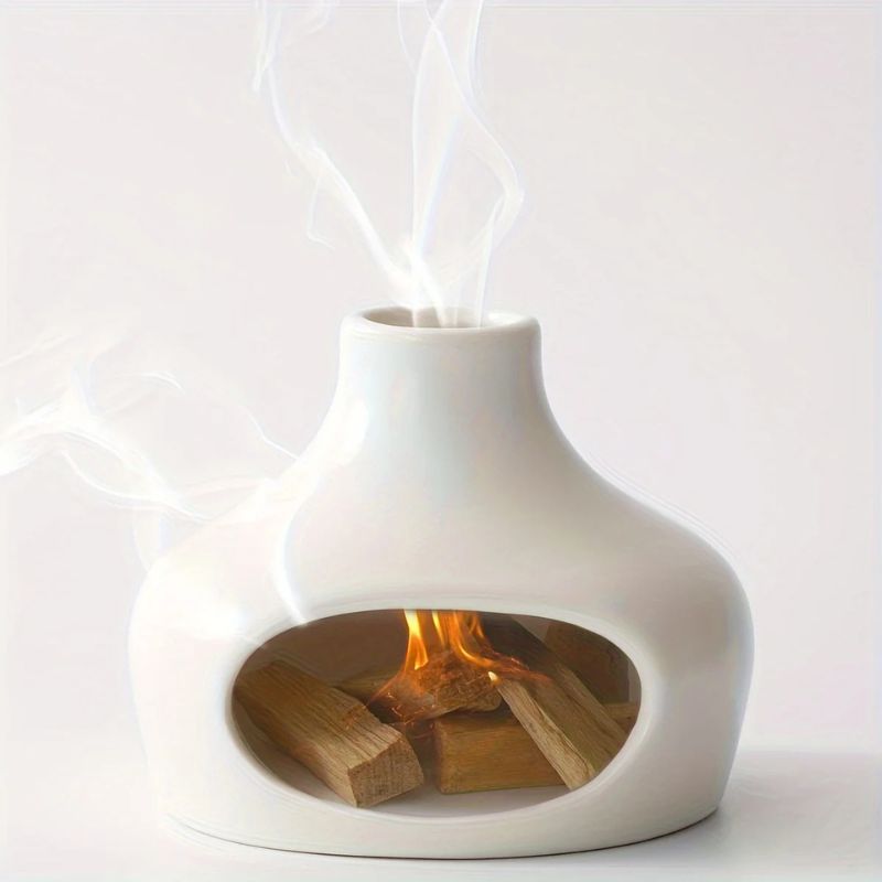 A white ceramic incense burner with a rounded shape and a narrow top opening, emitting smoke. Inside, wooden incense pieces are burning with a small flame visible through the front cutout.