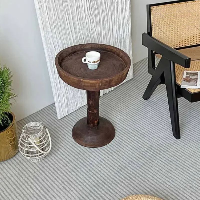 A rustic, dark wood, round side table (Momo Wabi-sabi Log Small Coffee Table) with a shallow tray top, holding a small cup, positioned on a striped rug with other decor elements, including a black chair with a woven back.