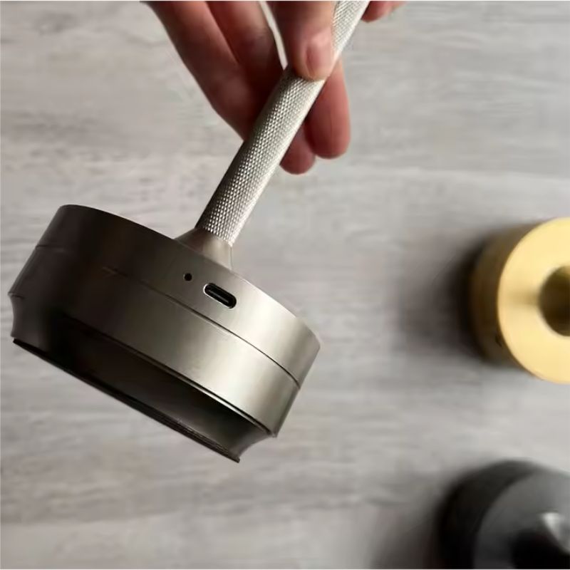 A close-up of a person holding a sleek, metallic lamp by its textured handle, showing the base with a charging port. Two additional lamp bases, one gold and one black, are visible on a wooden surface in the background.