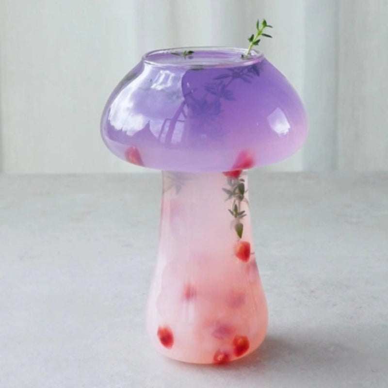 A clear glass cocktail glass in the shape of a mushroom sits on a light gray surface. The cap of the mushroom is filled with a translucent purple liquid and garnished with small green sprigs. The stem of the glass contains a lighter pink liquid with scattered red seeds or small fruit. The glass reflects light, highlighting its unique shape.