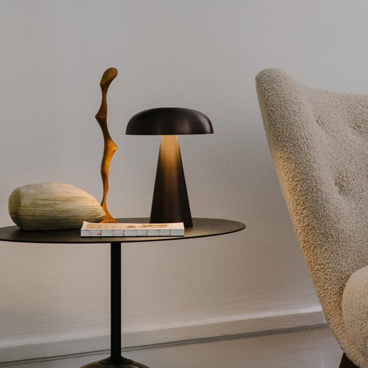 A cordless rechargeable table lamp with a dark, mushroom-shaped shade and a conical base emits a warm glow on a round black table. Next to it are a textured, light-colored stone, a slender wooden sculpture, and a small stack of white cards or papers. The arm of a textured beige chair is partially visible on the right. The scene is set against a light gray wall.