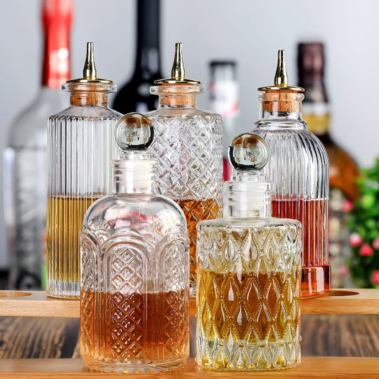 A collection of six clear glass bitters bottles with varied decorative patterns, some filled with amber liquid and featuring gold-toned dasher tops with cork stoppers, arranged on a wooden surface.