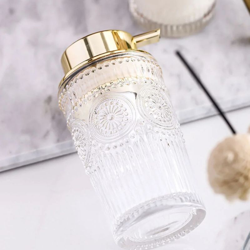 Ornate glass soap dispenser with a gold pump, angled on a marble surface, with a partial view of another dispenser and a decorative object in the background.