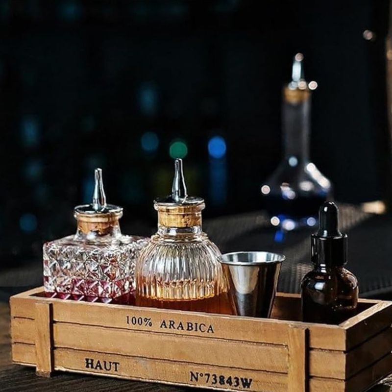 A wooden crate labeled "100% Arabica" holds two decorative glass bottles with spouts, a small metal jigger, and a dark dropper bottle, set against a dimly lit bar background.