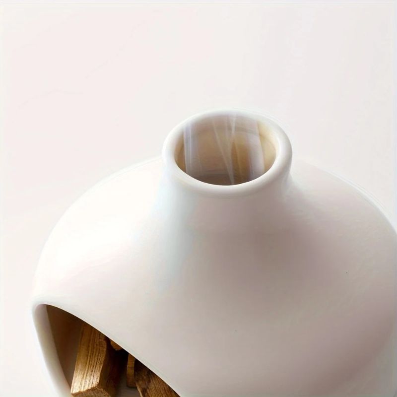 A close-up of a white ceramic incense burner with a rounded shape, a narrow top opening emitting smoke, and a front cutout revealing wooden incense pieces inside. The surface is smooth and glossy.