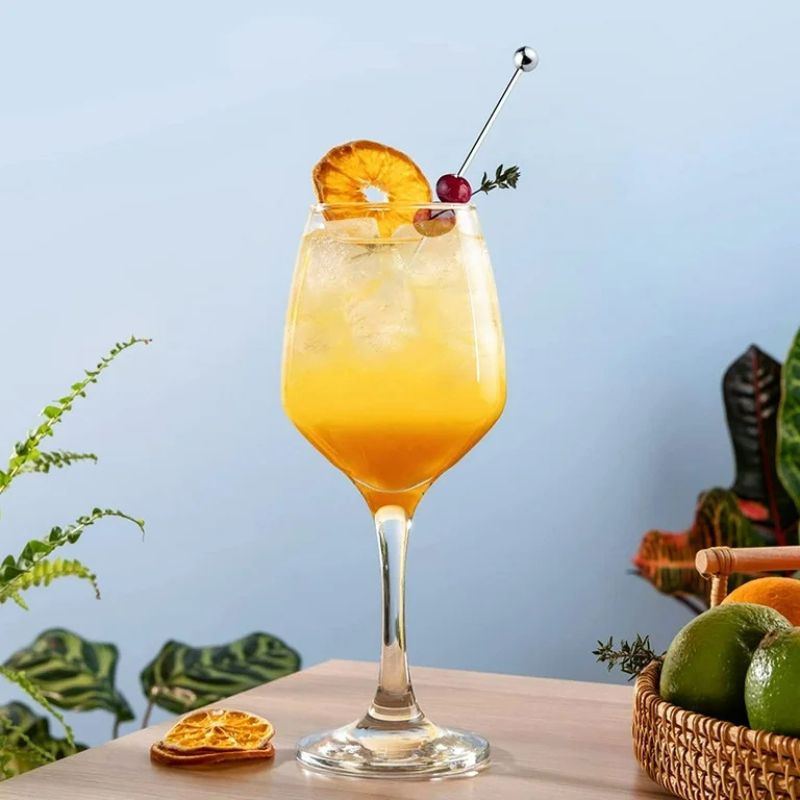 Orange cocktail in a stemmed glass with a dried orange slice, cherry, and ball-end cocktail pick, on a table with fruit and plants.